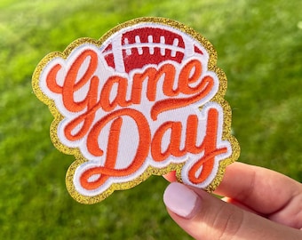 May include: A gold glitter iron-on patch with the words "Game Day" in orange and white lettering. A red and white football is above the text.