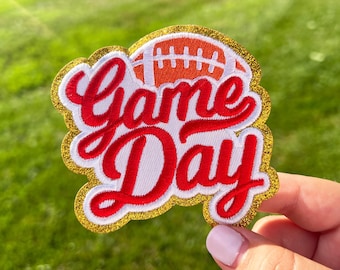 May include: A red and white embroidered patch with gold glitter trim. The patch features a football and the text "Game Day!"