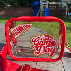 May include: Red clear plastic bag with a gold zipper and a red adjustable strap. The bag has two patches on it that say "Game Day" in red and gold letters.