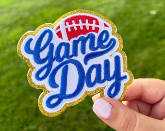 May include: A gold glitter iron-on patch with the words "Game Day" in blue and white lettering. A red and white football is above the words.