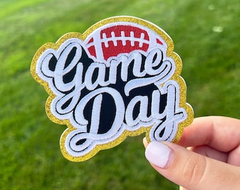 May include: A gold glitter iron-on patch with the words "Game Day" in white and black lettering. A red and white football is in the background.