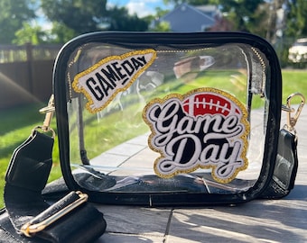 May include: Clear stadium-approved crossbody bag with black trim and gold hardware. Features two embroidered patches with the words "Game Day" and a football design. Includes a black adjustable strap.