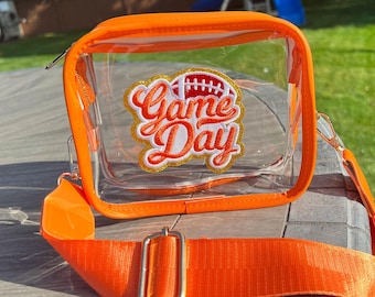 May include: Clear plastic crossbody bag with an orange trim and a strap. The bag has a patch on the front that says "Game Day" with a football graphic.