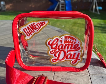 May include: Red clear plastic bag with a gold zipper and a red adjustable strap. The bag has two patches on it that say "Game Day" in red and gold letters.