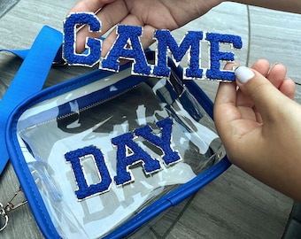May include: A clear, rectangular crossbody bag with a blue strap and trim. The bag has a zipper closure and the words "GAME DAY" in blue, textured letters with gold trim.