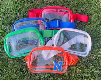 May include: Five clear plastic fanny packs with different colored trim. The packs are arranged in a semi-circle on a green grassy surface. The packs have adjustable straps and are clear enough to see through. One pack has a blue letter "N" on the front.