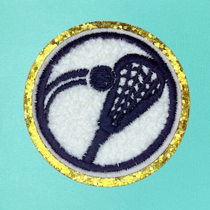 May include: A black and white embroidered lacrosse stick patch with a gold glitter border.