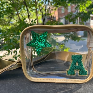 May include: Clear plastic crossbody bag with a gold trim. The bag has a green sequin star and a green felt letter "A" attached to the front.