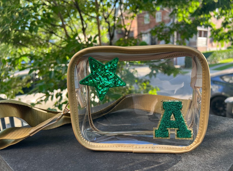 May include: Clear plastic crossbody bag with a gold trim. The bag has a green sequin star and a green felt letter "A" attached to the front.