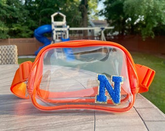 May include: A clear orange fanny pack with a blue felt letter "N" on the front. The fanny pack is made of clear plastic with an orange trim.