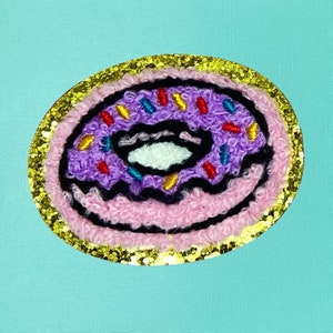 May include: A purple and pink donut with colorful sprinkles, embroidered on a pink fabric patch with a gold glitter border.