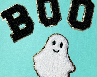 May include: Three black varsity-style letter patches spelling the word BOO, each featuring a gold glitter border. Below the letters is a white ghost patch with a textured surface, black embroidered eyes and mouth, and a gold glitter border.