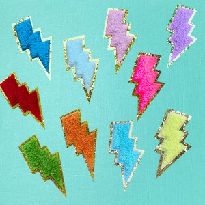 May include: Ten colorful lightning bolt shaped patches with gold glitter trim. The patches are made of fuzzy fabric and are in the colors red, orange, yellow, green, blue, light blue, pink, purple, and light purple.