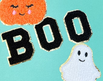 May include: A set of chenille patches featuring an orange pumpkin with a smiling face, the black letters B, O, and O, and a white ghost with a smiling face. Each patch has a gold glitter border and a textured, fuzzy surface finish.