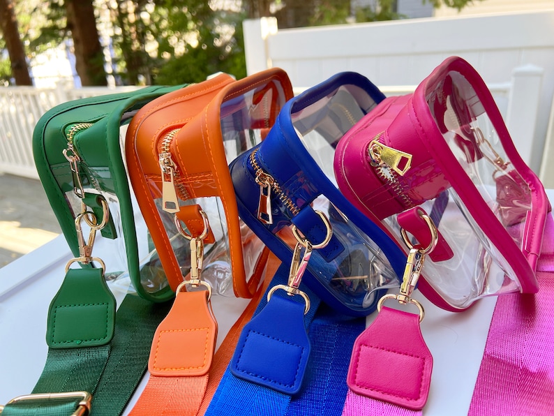 May include: Four clear plastic crossbody bags with colorful straps. The bags have gold zippers and gold hardware. The straps are green, orange, blue, and pink.