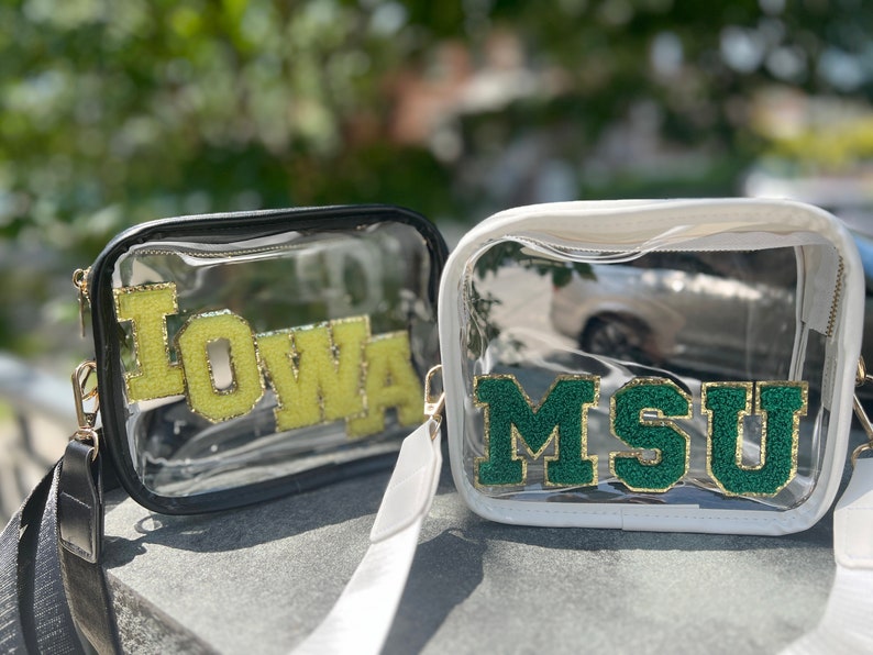 May include: Two clear plastic tote bags with black and white straps. The bag on the left has a yellow "IOWA" patch and the bag on the right has a green "MSU" patch.