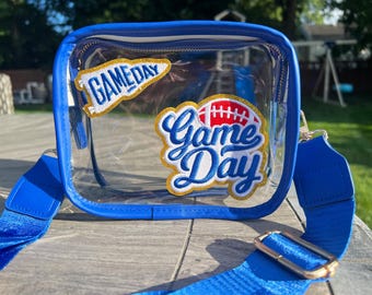 May include: A clear blue and white crossbody bag with a clear plastic front. The bag has two patches on the front, one that says "Game Day" with a football and the other that says "Game Day" in a banner shape.