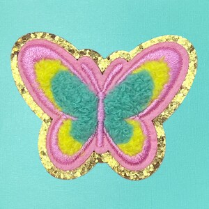 May include: A pink butterfly with yellow and green accents is embroidered on a gold glitter fabric. The butterfly is surrounded by a gold glitter border.