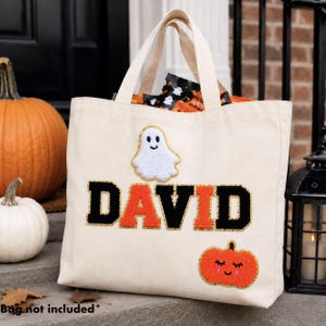 May include: A cream-colored canvas tote bag featuring the name DAVID in large, alternating black and orange varsity-style letters. Above the name is a white ghost patch, and below is an orange pumpkin patch with a smiling face.
