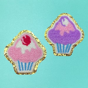 May include: Two embroidered cupcake patches with gold glitter trim. One cupcake is pink with white frosting and a red cherry on top. The other cupcake is purple with pink frosting.