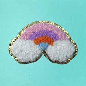 May include: A colorful rainbow-shaped patch with a gold glitter border. The rainbow features layers of pink, purple, orange, and blue, with fluffy white clouds. The patch is set against a light teal background.