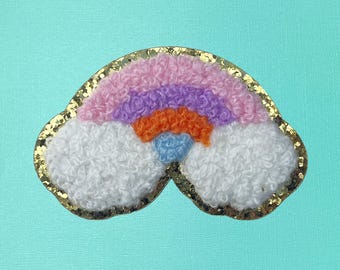 May include: A colorful rainbow-shaped patch with a gold glitter border. The rainbow features layers of pink, purple, orange, and blue, with fluffy white clouds. The patch is set against a light teal background.
