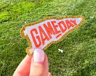 May include: A white felt pennant with gold glitter trim and the words "GAME DAY" embroidered in orange.