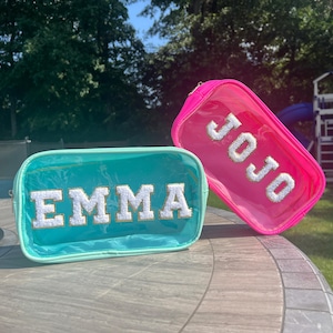May include: Two clear plastic zippered pouches with personalized names. The pouch on the left is teal blue with the name "EMMA" in white letters. The pouch on the right is pink with the name "JOJO" in white letters.