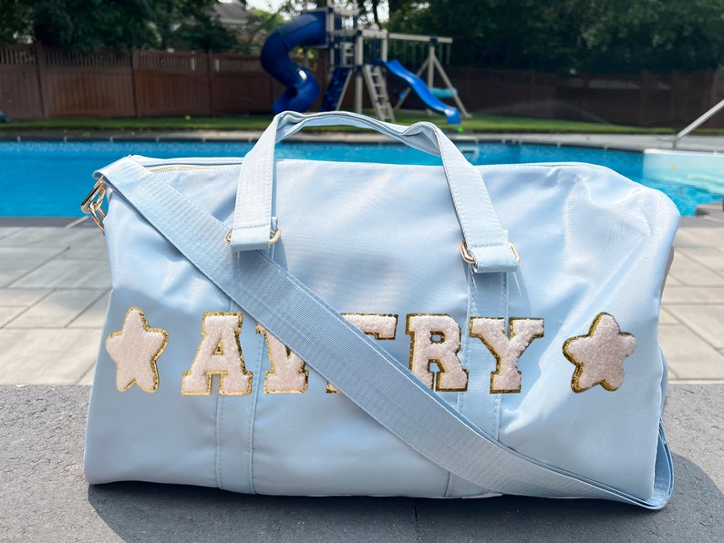 May include: Light blue duffel bag with a gold zipper and a white star on each side. The bag has the name "AVRY" in gold letters with a white outline.