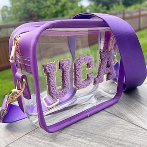 May include: A clear purple and gold crossbody bag with a zipper closure and a clear front panel. The bag has the letters "UCA" in fuzzy purple letters with gold glitter on the front.