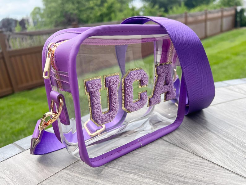 May include: A clear purple and gold crossbody bag with a zipper closure and a clear front panel. The bag has the letters "UCA" in fuzzy purple letters with gold glitter on the front.