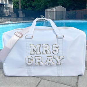 May include: White travel duffel bag with a gold zipper and a white strap. The bag has the text "MRS GRAY" in gold letters on the front.