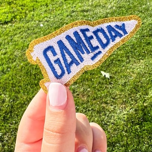 May include: A white and blue pennant-shaped patch with the word "GAMEDAY" in blue block letters. The patch has a gold glitter border and is set against a background of green grass.