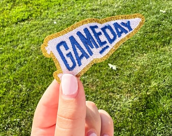 May include: A white and blue pennant-shaped patch with the word "GAMEDAY" in blue block letters. The patch has a gold glitter border and is set against a background of green grass.