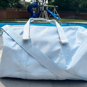 May include: A light blue duffel bag with a gold zipper and gold hardware. The bag has a long strap that can be worn over the shoulder.
