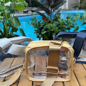 May include: Three clear plastic crossbody bags with different colored straps. The bags are on a wooden surface. The straps are gold, silver, and navy blue.