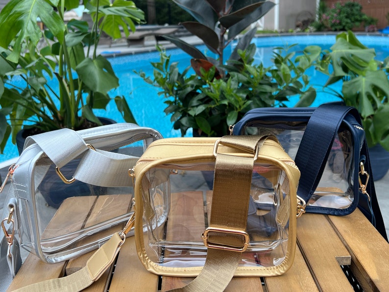 May include: Three clear plastic crossbody bags with different colored straps. The bags are on a wooden surface. The straps are gold, silver, and navy blue.