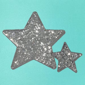 May include: Two silver glitter star-shaped patches on a turquoise background. The larger star is on top of the smaller star.