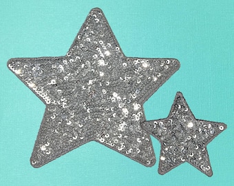 May include: Two silver glitter star-shaped patches on a turquoise background. The larger star is on top of the smaller star.
