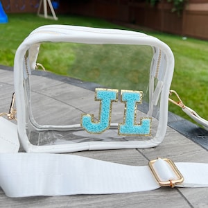 May include: A clear plastic crossbody bag with a white strap and a gold buckle. The bag has a white zipper and a white trim. The bag has the letters "JL" in blue and gold glitter on the front.