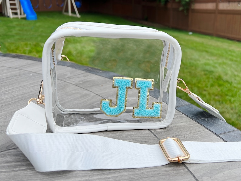 May include: A clear plastic crossbody bag with a white strap and a gold buckle. The bag has a white zipper and a white trim. The bag has the letters "JL" in blue and gold glitter on the front.