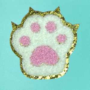 May include: A white and pink paw print with gold glitter around the edges. The paw print is made of fluffy fabric.
