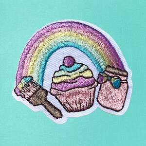 May include: Embroidered iron-on patch featuring a rainbow, cupcake, paintbrush, and jar. The rainbow is in shades of pink, yellow, and blue. The cupcake is pink, yellow, and purple with a blue swirl on top. The paintbrush is brown with blue paint. The jar is pink with a blue lid and a blue bow.