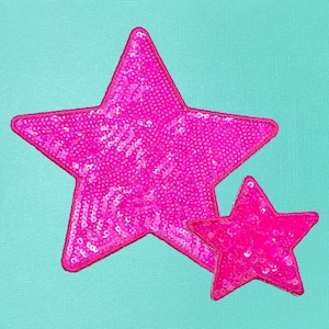 May include: Two pink sequined star patches on a teal background. The larger star is positioned above and to the left of the smaller star. The stars have a stitched outline.