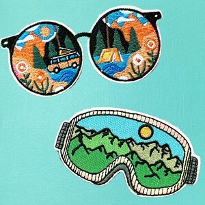 May include: Embroidered patches featuring outdoor scenes. One patch depicts sunglasses with a camping scene inside the lenses. The other patch shows ski goggles with a mountain range and sun. Patches are in shades of orange, blue, green, and black.