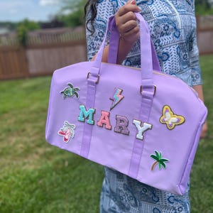 Puede incluir: Una bolsa de lona de color lavanda con cierre de cremallera y herrajes dorados. La bolsa tiene varios parches coloridos, incluyendo una tortuga, un rayo, el nombre "MARY", una mariposa y una palmera.