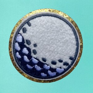 May include: A white golf ball with a black and blue embroidered design, surrounded by a gold glitter border.