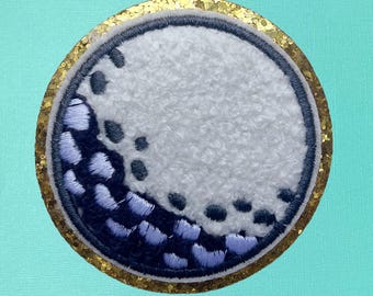 May include: A white golf ball with a black and blue embroidered design, surrounded by a gold glitter border.