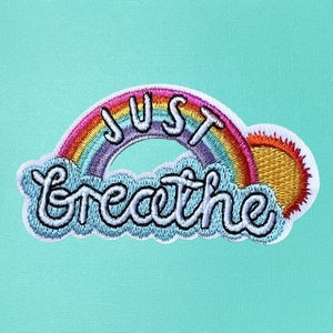 May include: A white embroidered patch with a rainbow arch and the words "JUST breathe" in a white cursive font. The rainbow has red, orange, yellow, green, blue, indigo, and violet stripes. The sun is yellow with orange rays.