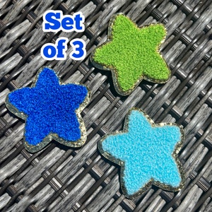 May include: Set of three star-shaped fabric patches. The stars are blue, green, and light blue with gold glitter trim.  "Set of 3" is written in blue text on a white background.